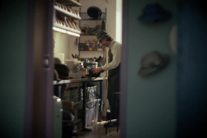 An artisan hatter carefully crafts a hat on a sewing machine in his sunlit workshop, viewed through a doorway