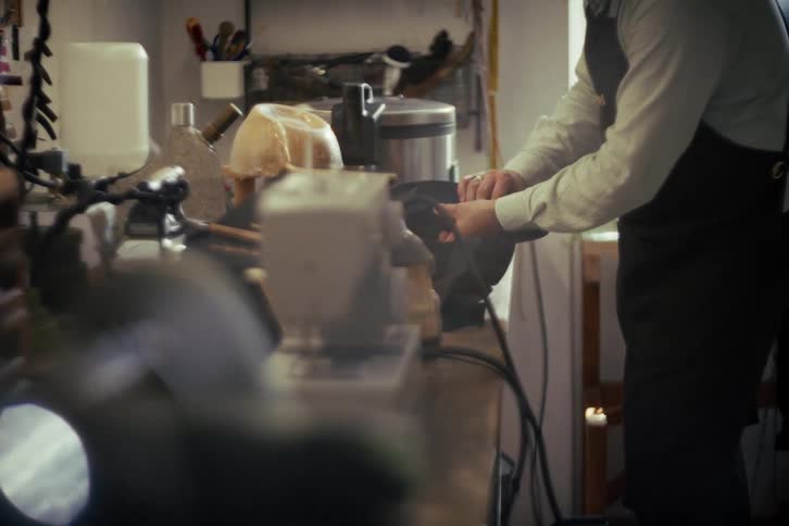 An artisan in a leather apron meticulously works on a shoe held in a vise in a traditional, sunlit workshop