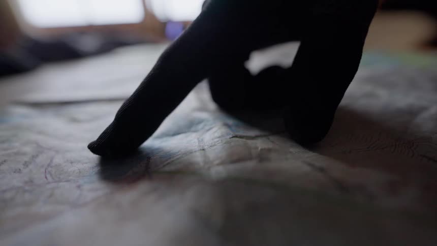 A gloved finger slowly traces a route on a weathered topographical map laid out on a table