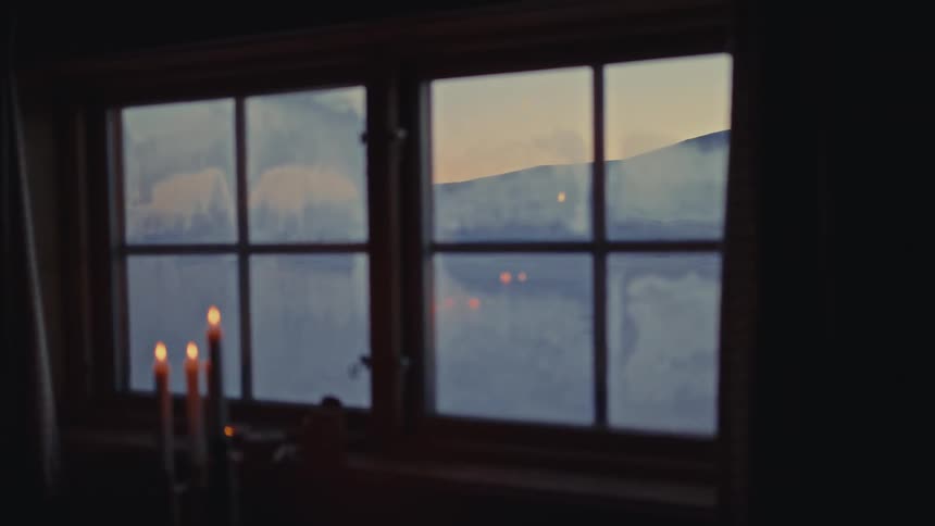 A soft-focus view from a dark room with lit candles on a windowsill looking out at a snowy landscape during twilight