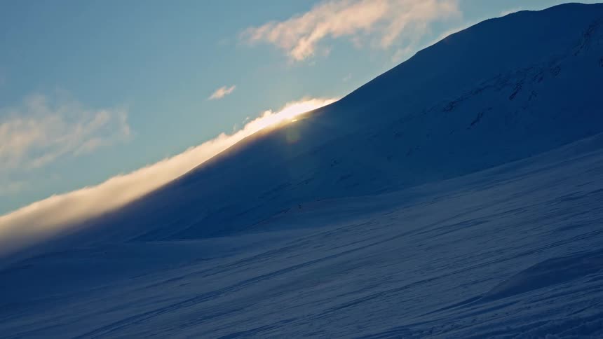 Sunlight illuminates clouds moving over a vast, snowy mountain slope in a peaceful cinemagraph