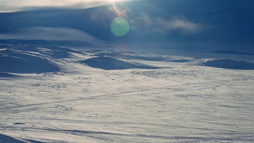 A vast and desolate frozen landscape is illuminated by sunlight streaming through low clouds and mist, creating a bright lens flare