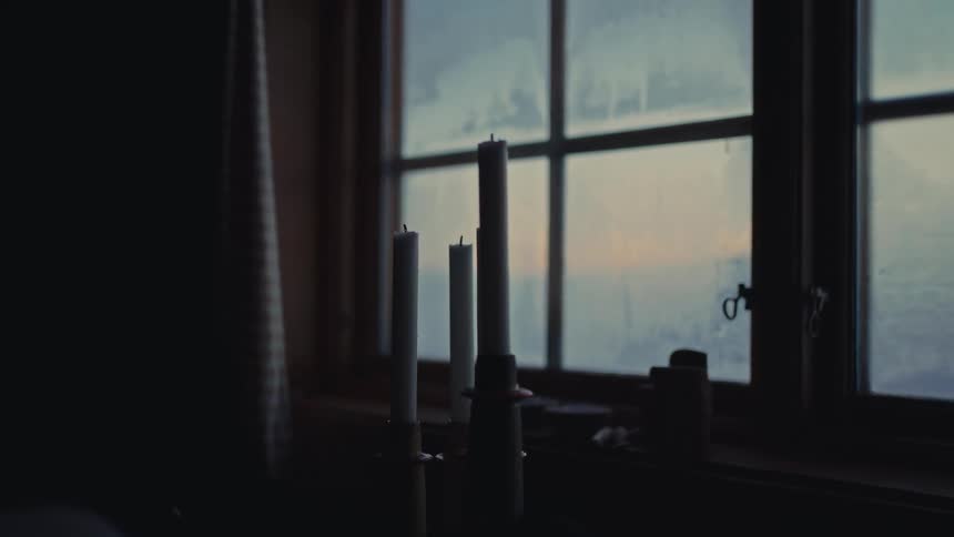 Unlit candles sit silhouetted on a windowsill in a dark room as soft, hazy light filters through the window