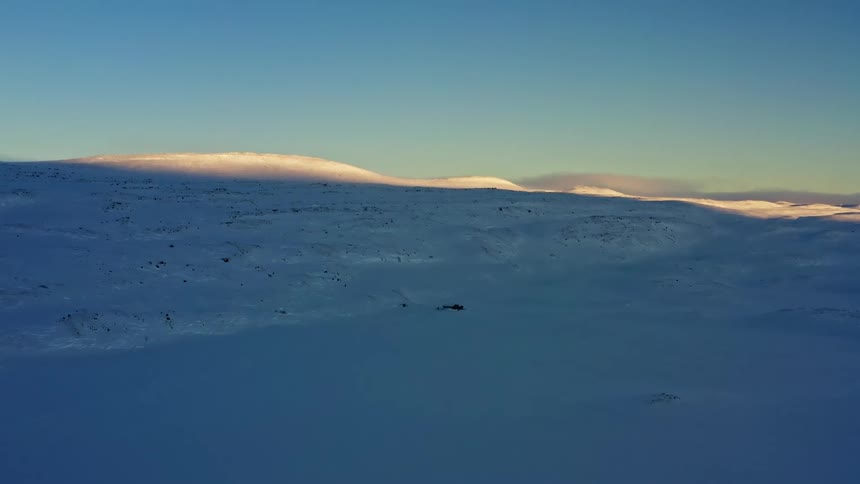 A tranquil, wide, static shot of a vast, snow-covered tundra with golden sunrise light touching the distant mountain peaks
