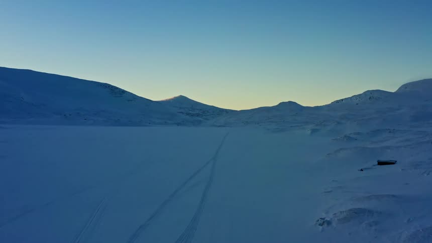 An aerial view slowly advances over a vast, frozen landscape with snowmobile tracks disappearing into the distance towards mountains at dawn