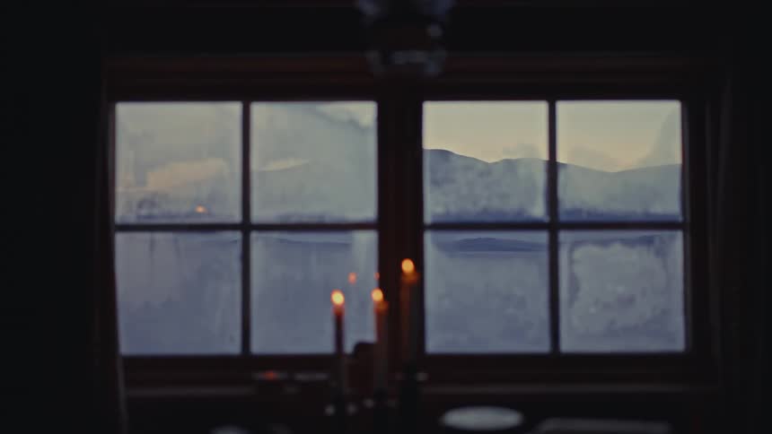 A serene view of a mountain landscape is seen through a frosted window from a dark, candlelit room
