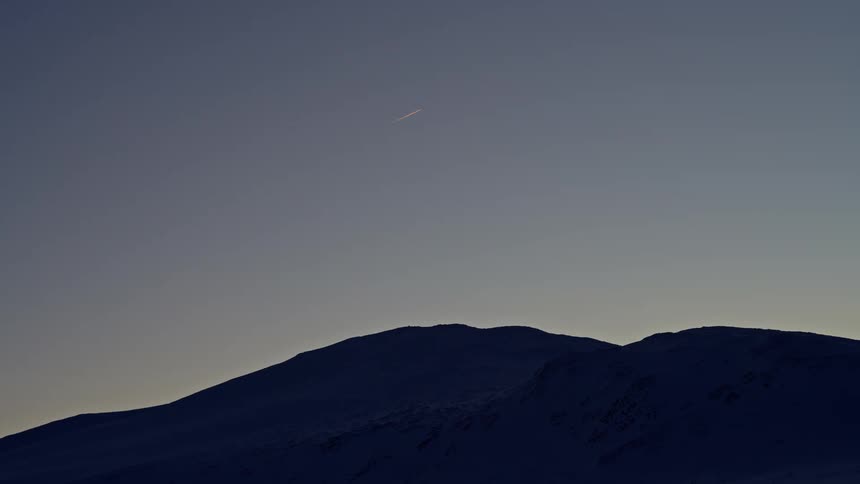 A shooting star streaks across the tranquil twilight sky above a silhouetted, snow-capped mountain