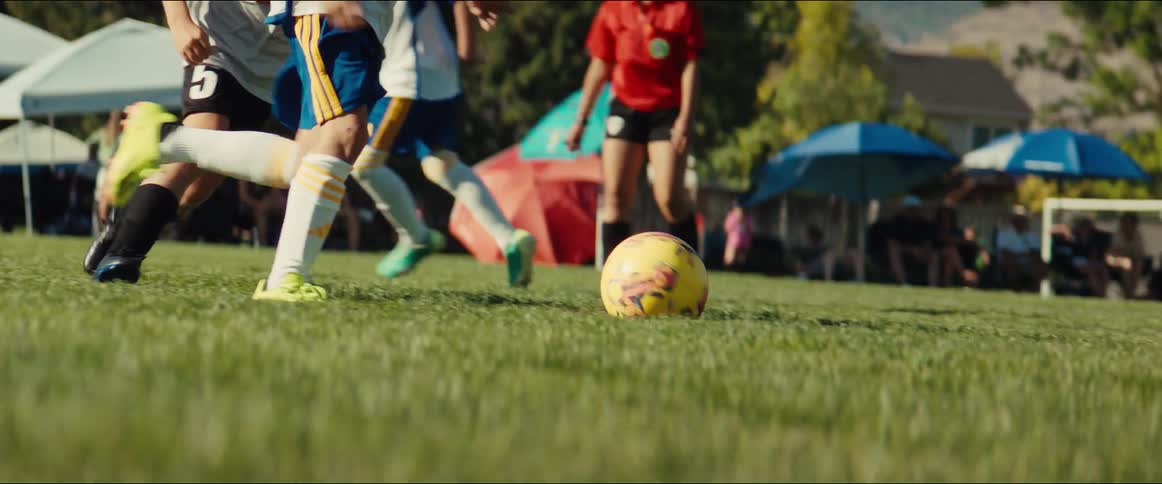 A low-angle, slow-motion shot follows the action as young boys run and kick a soccer ball down the field during a game