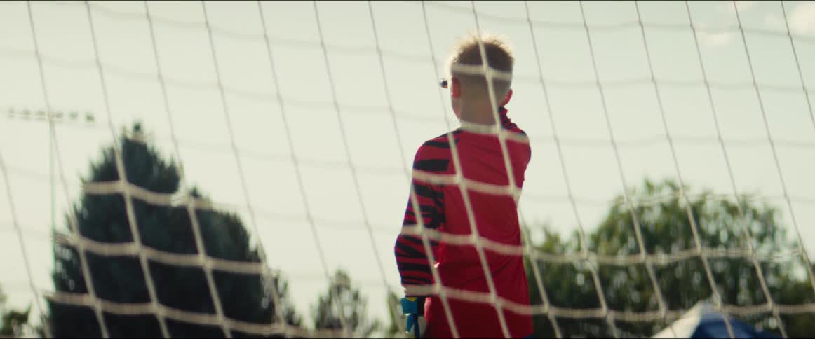 A young boy in a red goalie uniform stands inside a soccer net, waiting for the game on a sunny day
