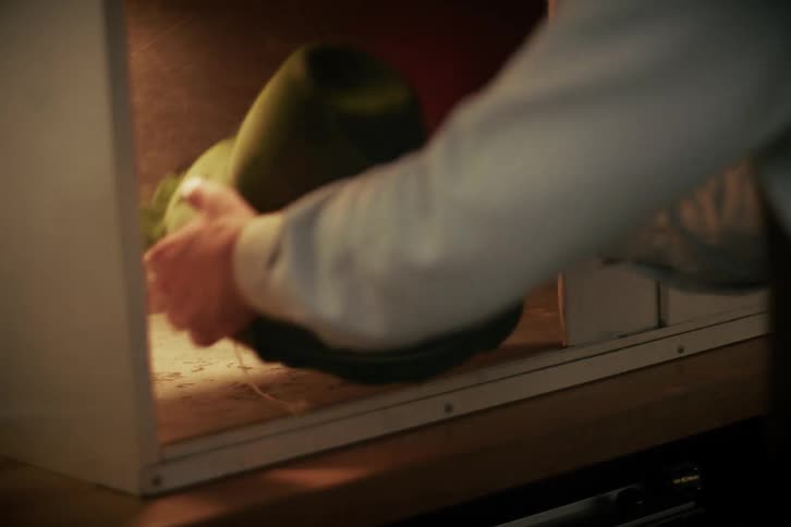 An artisan slides open a lit cabinet and carefully places a green felt hat inside before closing the door
