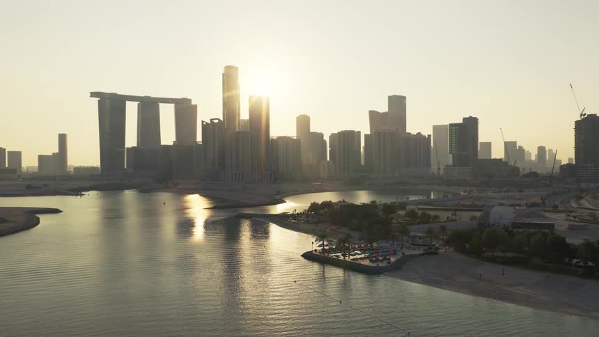 Aerial view of the modern Abu Dhabi skyline with its iconic Gate Towers reflecting in the calm ocean water during a golden sunrise
