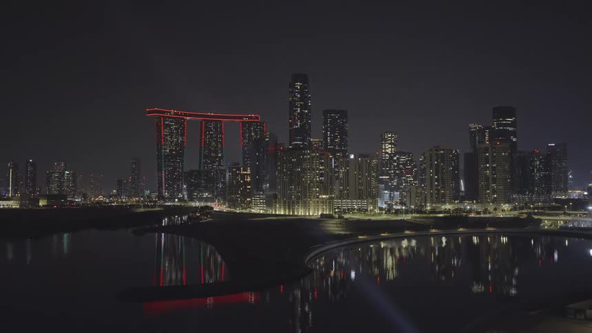 A stunning aerial view of the illuminated Al Reem Island cityscape in Abu Dhabi at night, reflecting in the calm water below