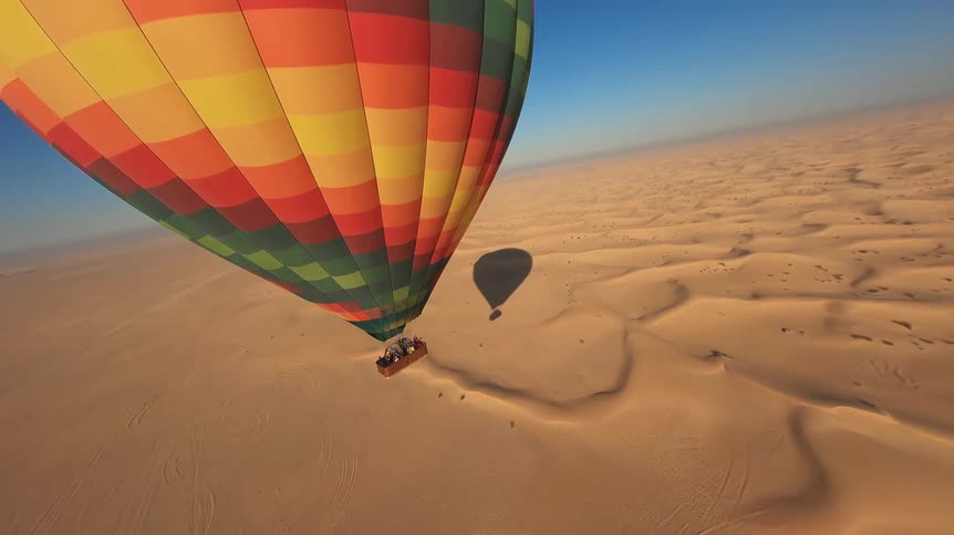 A colorful hot air balloon flies low over the vast, sunlit sand dunes of a desert