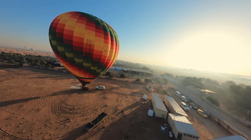 Descending aerial view of a vibrant, multi-colored hot air balloon being prepared for flight in the desert at sunrise