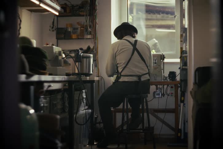 A traditional hatter sits at a workbench in his workshop, carefully crafting a new felt hat