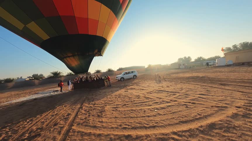 A colorful hot air balloon lifts off from the desert sand as the sun rises, with crew and passengers preparing for the flight
