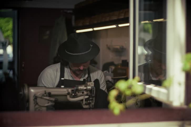A skilled artisan wearing a fedora hat carefully works on a vintage sewing machine in his dimly lit workshop, as seen from an outside window