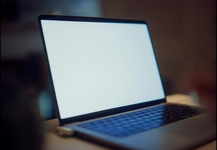 A modern laptop with a blank white screen sits on a desk in a dimly lit room, perfect for screen replacement content
