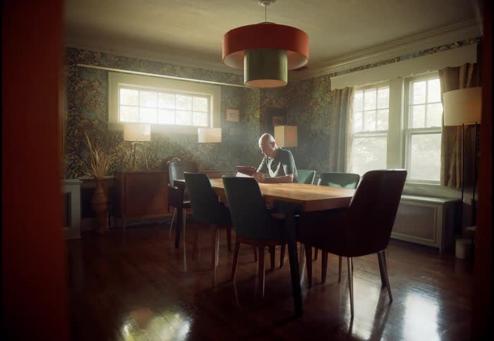 An elderly man sits alone at a long dining table in a dramatically lit room, reading quietly to himself