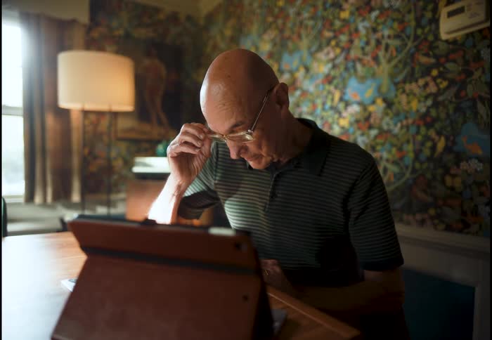 An elderly man in a dimly lit room takes off his glasses to look closely at his tablet computer