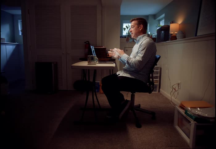 A man working from home in a dimly lit basement office engages in a video call on his laptop, gesturing as he speaks