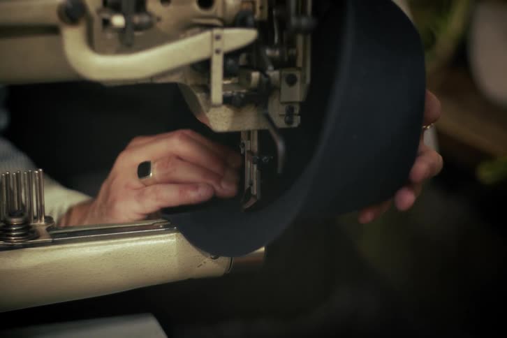 A close-up shot of an artisan's hands skillfully using an industrial sewing machine to stitch a dark felt hat in a workshop
