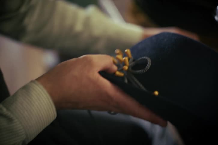A craftsman's hands meticulously hand-sew a decorative cord with gold tassels onto a dark blue felt hat