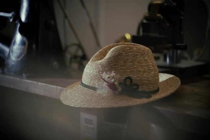 A static, close-up shot of a stylish straw hat with a feather accent resting on a wooden table in a vintage workshop