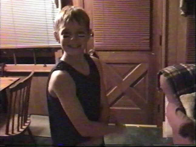 A playful young boy with football makeup on his face smiles and waves excitedly for the camera in his home