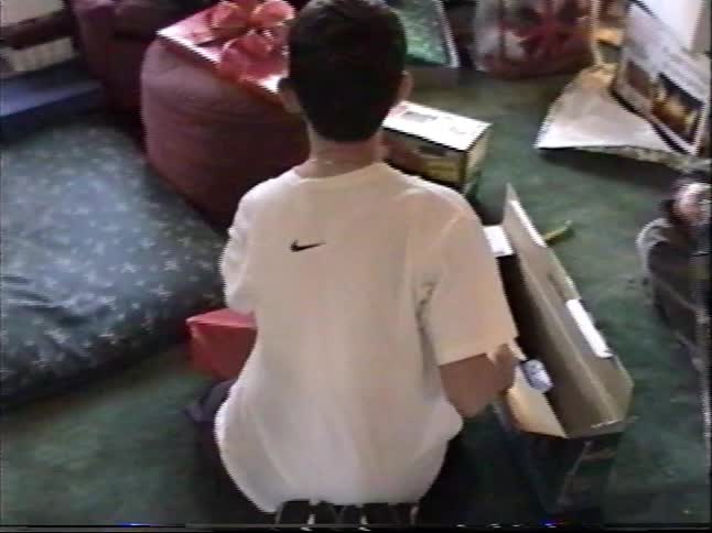 Vintage home video captures a boy from behind opening presents with his family on Christmas morning in the 90s