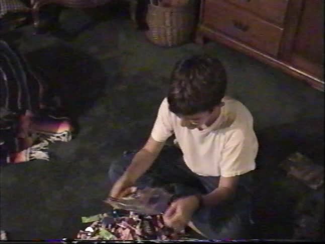 A young boy sits on a dark green carpet and pours a large bag of assorted candy onto the floor in this nostalgic home video
