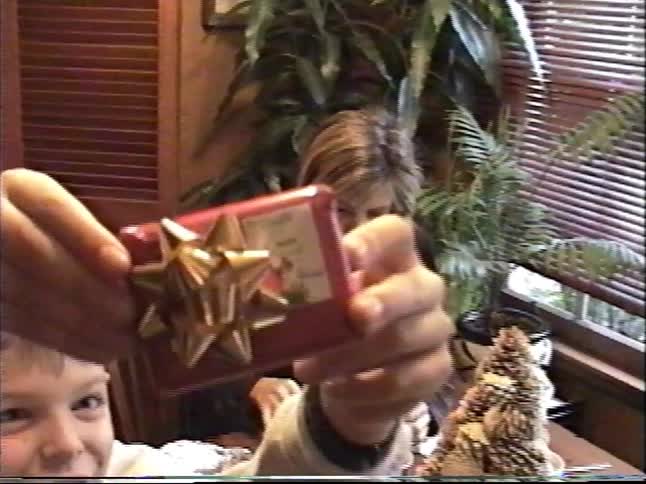 A young boy holds a small Christmas present up to the camera while his mother eats at the dining table in this vintage home video