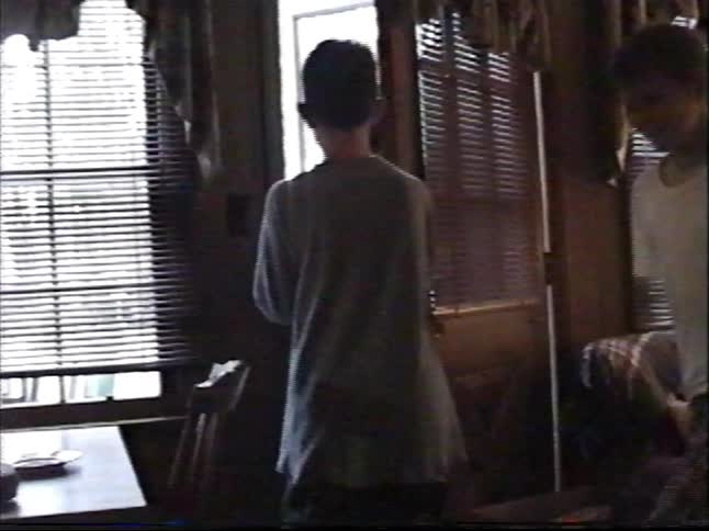 A vintage home video shows young men in a living room, one opening a door and another bending over laughing