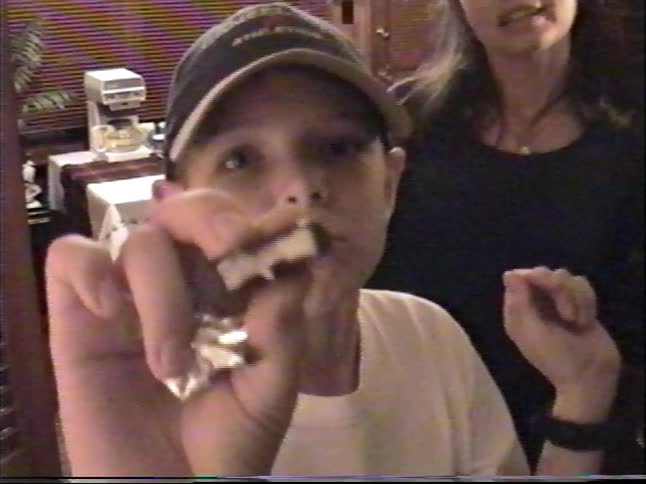 Vintage home video footage shows a woman talking in a doorway as a young boy eats a chocolate candy and shows it to the camera