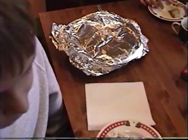 A young boy smiles while enjoying a cozy Christmas breakfast with his family, featuring warm cinnamon rolls and orange juice, captured on nostalgic home video