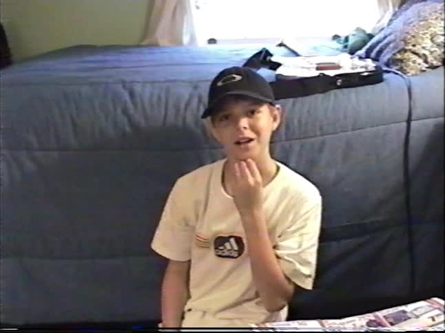A thoughtful young boy in a baseball cap talks to the camera while sitting on the floor of his messy bedroom in this vintage home video