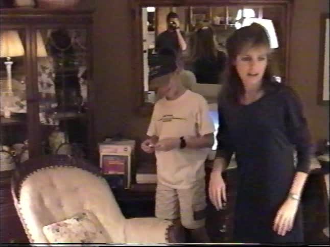 A candid family moment from the 90s shows a young boy in a baseball cap proudly showing a small rock to his dad filming on a camcorder