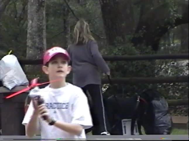 Vintage home video footage shows a young boy launching a toy propeller helicopter into the air at a park