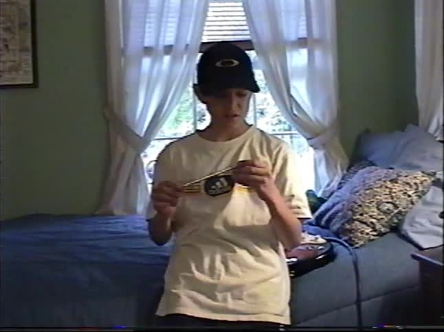 A young boy in a baseball cap plays with a pencil in his bedroom before sitting on the floor to write on a piece of paper