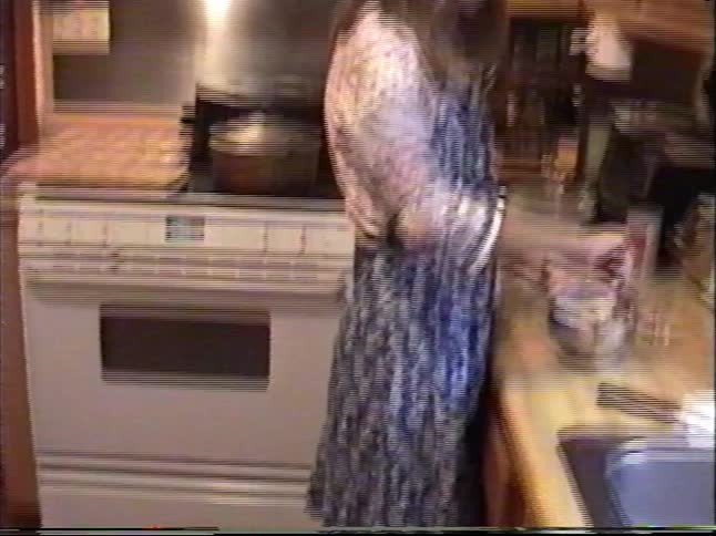 A woman cooks in a cozy home kitchen before the camera pans down to her black lab resting on the floor in this vintage VHS home movie
