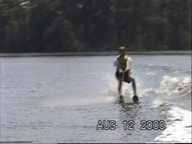 Vintage home video footage shows a man water skiing across a calm lake on a summer day in the year 2000