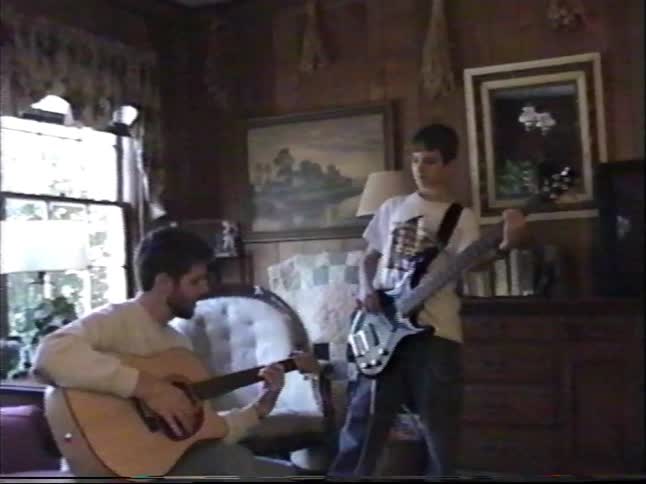 Friends and family of varying ages jam on acoustic and bass guitars in a cozy living room, captured on vintage home video