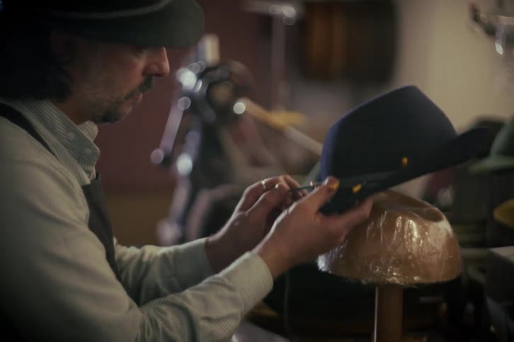 An artisan milliner carefully sews the final details onto a custom-made felt hat in his traditional workshop