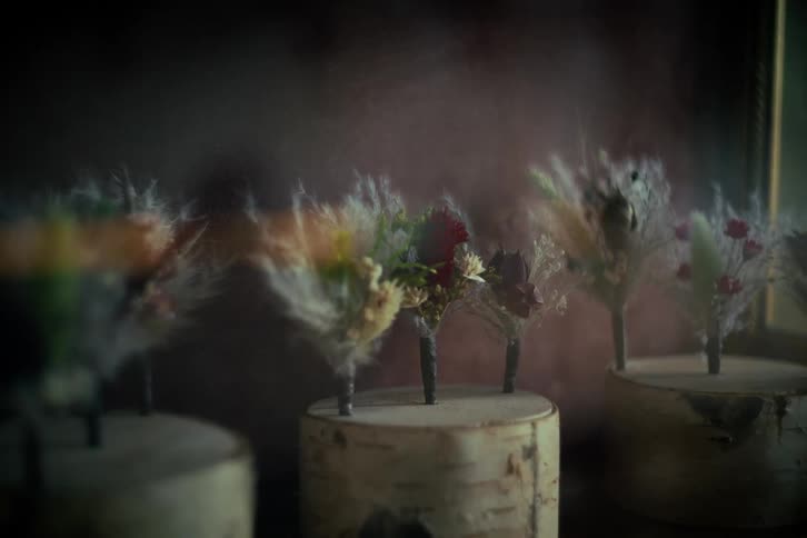 A fine mist is sprayed over rustic wedding boutonnieres made of dried flowers and grasses, resting on birch wood stands