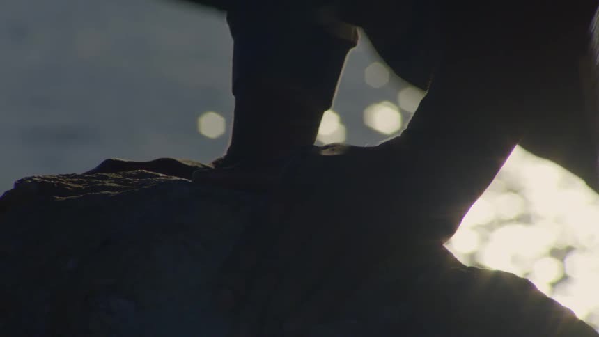Close up silhouette of a person's feet standing on a rock overlooking shimmering water