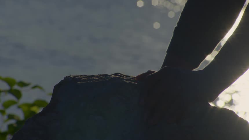 The silhouette of a person's hands rests peacefully on a large rock by a shimmering lake during golden hour