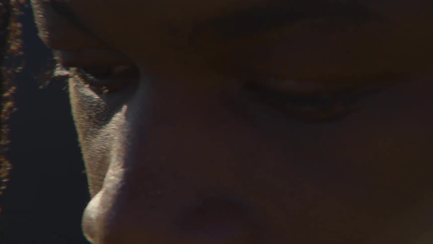 An intimate close-up shot focuses on the pensive face of a person with dreadlocks, their eyes cast downward in deep thought