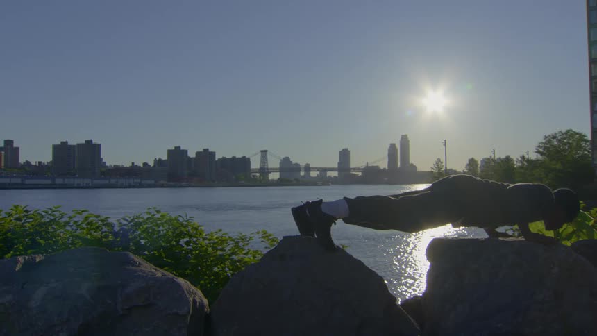 A silhouetted man performs inclined push-ups on rocks by the river with the city skyline and bright sun in the background
