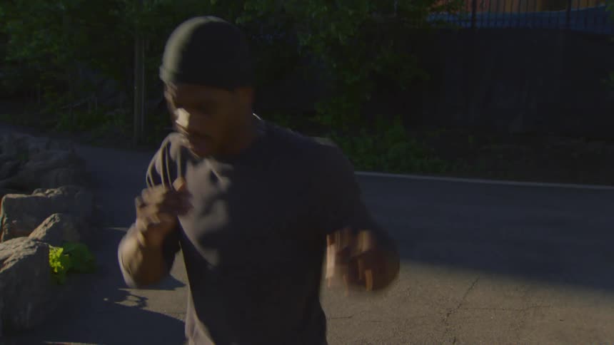 An athletic man in a black beanie and shirt warms up for his workout by stretching and bouncing on a park path during golden hour