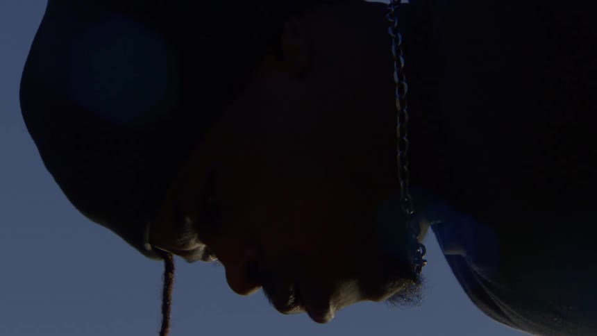 A low-angle close-up silhouette of a man in a hoodie with his head bowed against a clear blue sky